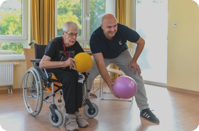 Therapeut macht mit einem Bewohner im Rollstuhl eine Ballübung im Tagesraum